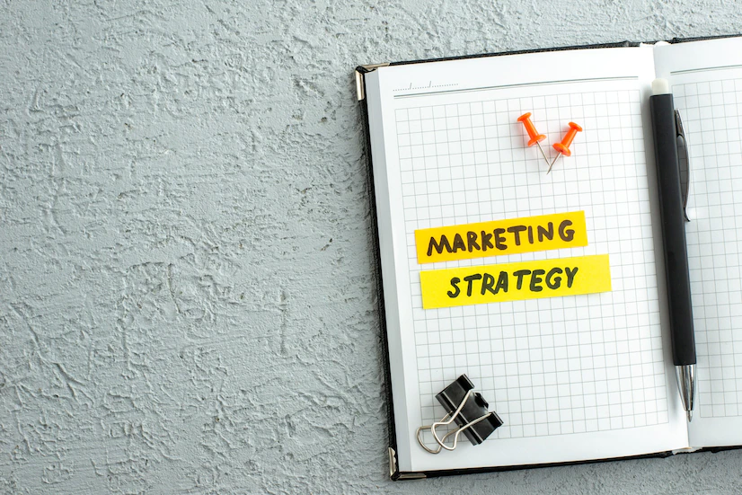 vista-superior-pluma-escritura-estrategia-marketing-cuaderno-espiral-abierto-sobre-fondo-arena-gris_179666-20009